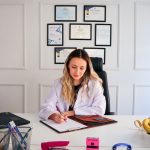 Nutritionist sits at a modern office desk writing, with fruits and diplomas displayed, emphasizing health and professionalism.